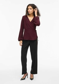 Burgunderfarbene Wickeloberteil mit langen Ärmeln und Bindedetail, kombiniert mit schwarzen weit geschnittenen Hosen und spitzen schwarzen Slingback-Pumps.