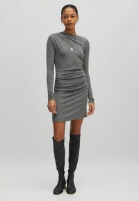 Graues Langarmkleid mit gerafften Details, figurnah geschnitten und aus weichem Material. Getragen mit schwarzen kniehohen Stiefeln. Schlichtes, akzentuiertes Halskettchen.