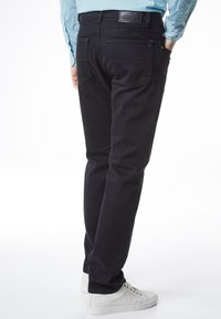 Hombre con pantalones vaqueros negros, camisa a cuadros azul y zapatillas blancas, de pie con las manos en los bolsillos, visto desde atrás.