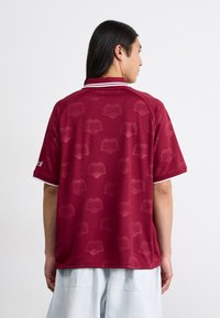 Burgundská polo košeľa s jemným vzorom srdca, bielym golierom a krátkymi rukávmi. Vyrobená z hladkej, ľahkej tkaniny.