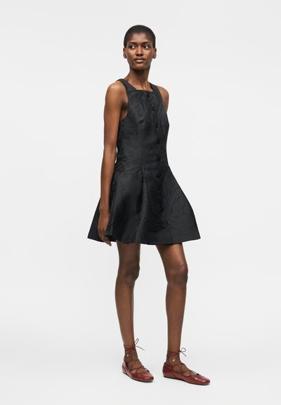 Γυναίκα που στέκεται, φορά μαύρο αμάνικο φόρεμα γραμμής Α με κουμπιά μπροστά και κόκκινα δετά φλατ παπούτσια, λευκό φόντο.