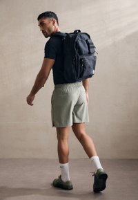 Sac à dos en tissu noir avec plusieurs poches et boucles latérales, porté par une personne en chemise noire ajustée et shorts verts clairs, s'éloignant.