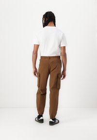Brune cargo bukser med elastiske manchetter, sidelommer og en kontrasterende talje, parret med en hvid t-shirt og sorte sneakers.