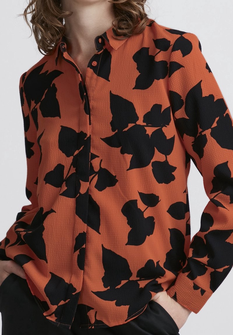 Camicia a maniche lunghe con bottone, realizzata in tessuto arancione con una superficie testurizzata, decorata con grandi motivi a foglia nera. Maniche lunghe con polsini a bottone.