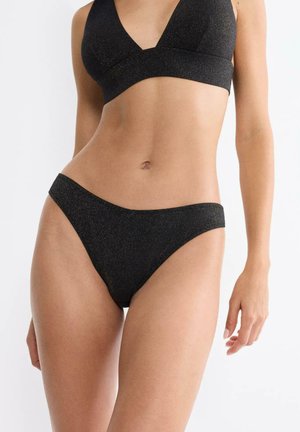 Kobieta w czarnym błyszczącym komplecie bikini, stojąca na tle jednolitego jasnego tła, pokazująca tułów i górną część nóg.