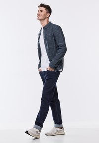 Outfit casual composto da una camicia a maniche lunghe blu scuro con texture sopra una t-shirt bianca, abbinata a jeans scuri e sneakers bianche.