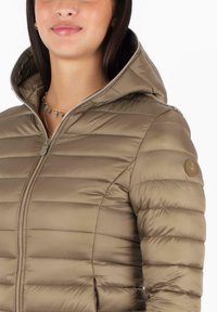 Giacca leggera e imbottita di colore beige con zip frontale, cappuccio e una toppa circolare con logo sulla manica sinistra. Texture liscia e trapuntata.