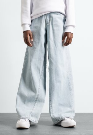 Person iført lyseblå brede jeans, hvide sneakers og en hvid langærmet top, stående på en grå gulv.