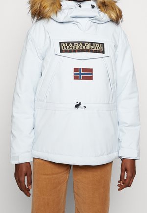 Veste d'hiver blanche avec capuche bordée de fourrure, poche avant, patch du drapeau de la Norvège et logo Napapijri sur la poitrine, portée avec un pantalon en velours côtelé beige.