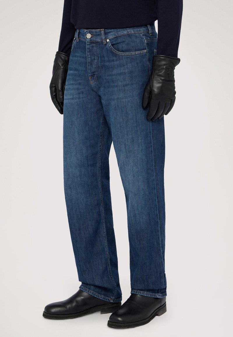 Jeans de mezclilla azul oscuro con un corte relajado, que presentan un diseño clásico de cinco bolsillos, remaches plateados, y se combinan con guantes y botas de cuero negras.