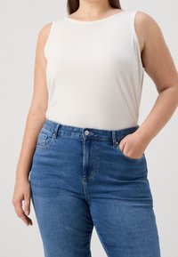 Persona vistiendo una camiseta sin mangas de color blanco con textura acanalada metida dentro de unos pantalones vaqueros de talle alto azules, con una mano en el bolsillo, contra un fondo liso.