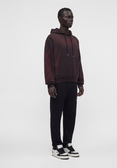 Homme portant un sweat à capuche bordeaux foncé avec texte rouge sur la manche et un pantalon noir, debout devant un fond gris clair uni.
