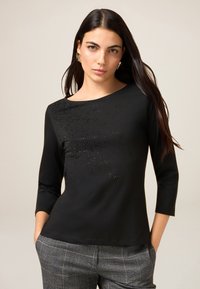 Sort langærmet top med bådudsk�æring og pyntet med spredte rhinsten foran. Blødt, elastisk stof.
