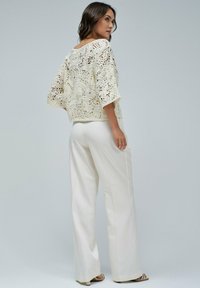 Lace-Strick-Croptop in Creme mit weitem Ausschnitt, 3/4-Ärmeln und aufwendigen floralen Mustern, kombiniert mit weißen weit geschnittenen Hosen.