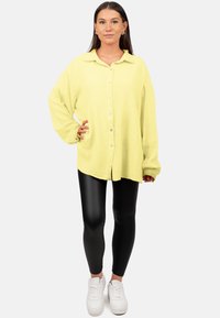 Camicia giallo limone oversized con bottoni in tessuto testurizzato, abbinata a leggings neri e sneakers bianche. Vestibilità casual e rilassata.