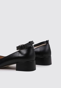Chaussures en cuir noir avec un petit talon carré, bout rond et une bride de cheville avec une boucle en métal. Texture lisse avec un design minimaliste.