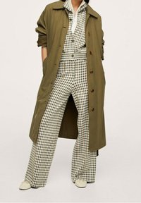Oliwkowy trench coat noszony na dopasowanym, w kratkę zestawie w białe i zielone. Szerokie spodnie mają teksturowany wzór. Białe buty dopełniają całość.