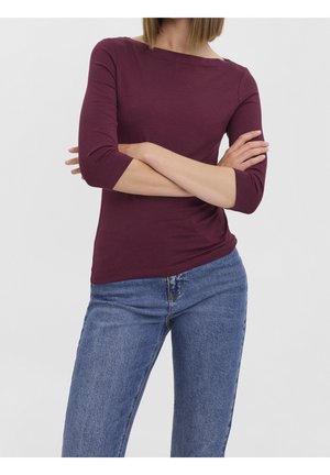 Vrouw met een bordeaux top met driekwart mouwen en een blauwe spijkerbroek, staand met gekruiste armen tegen een effen achtergrond.
