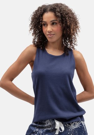 Mujer con cabello rizado que lleva una camiseta sin mangas azul marino y pantalones azul marino con estampado y cordones blancos sobre un fondo claro.