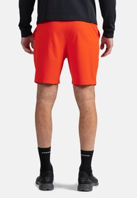 Pantaloni sportivi arancioni brillanti con vita elastica, texture liscia e tasche laterali, abbinati a calze e scarpe nere.