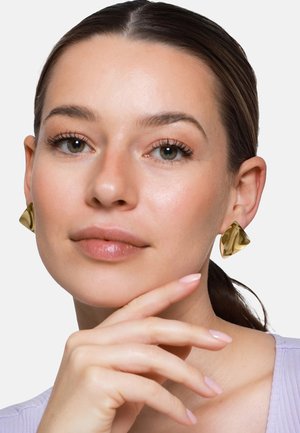 Kobieta o jasnej karnacji i ciemnych włosach, nosząca duże złote trójkątne kolczyki, opierająca podbródek na dłoni z paznokciami pomalowanymi na jasnoróżowy kolor.