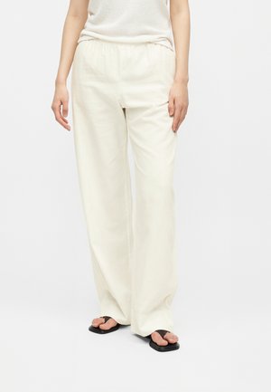 Osoba nosiaca voľné, vysoko v páse, krémové nohavice v kombinácii s čiernymi sandálmi, stojaca pred jednoduchým svetlým pozadím.