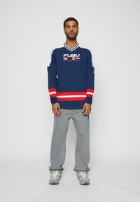 Cămașă cu mânecă lungă FUBU de culoare navy, cu accente roșii și albe în dungi, logo pe piept, asortată cu blugi gri și adidași gri.