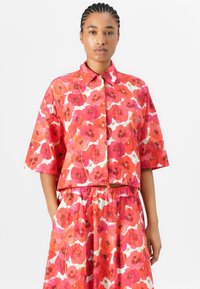Rotes, blumiges Cropped-Shirt mit kurzen Ärmeln, das Mohnblumenmuster auf weißem Hintergrund zeigt, kombiniert mit einem passenden Rock.
