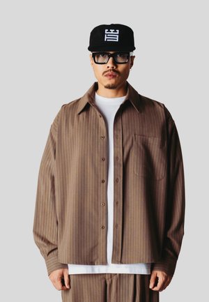 Homme portant un ensemble chemise et pantalon marron à fines rayures sur un t-shirt blanc, casquette noire avec logo blanc et grandes lunettes à monture noire.