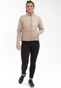 Beige isolerad jacka med hög krage, ribbad struktur på den övre framsidan, kombinerad med svarta åtsittande leggings och mörka sportskor.