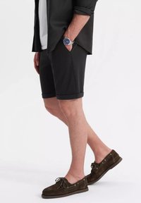 Schwarze Baumwollshorts mit hochgekrempelten Säumen, Seitentaschen, getragen mit einem grauen Shirt und braunen Wildlederbootsschuhen. Silberne Uhr am Handgelenk.