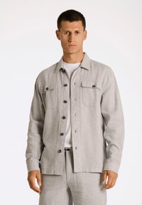 Grå knäppskjorta med två bröstfickor, tillverkad av texturerad tyg. Har svarta knappar och krage, bärs över en vit t-shirt.