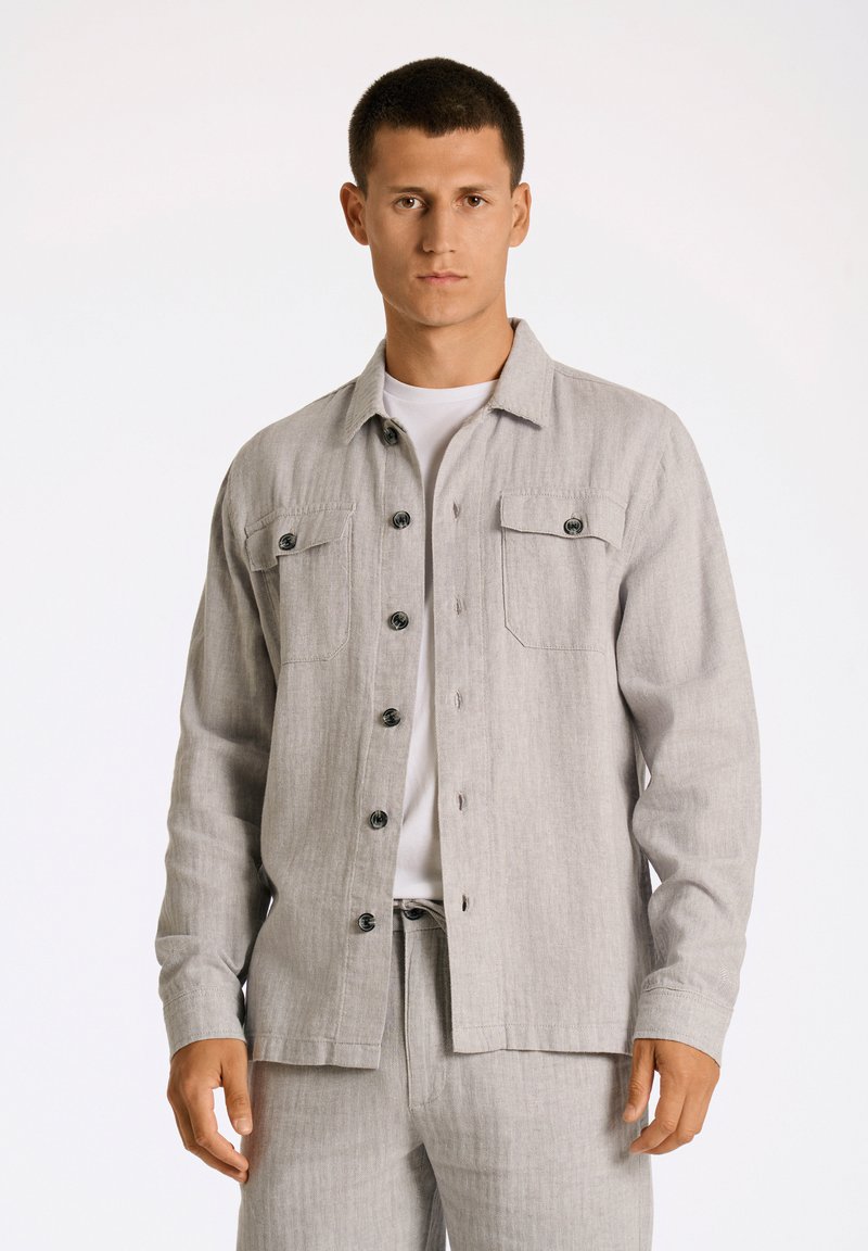 Grå knäppskjorta med två bröstfickor, tillverkad av texturerad tyg. Har svarta knappar och krage, bärs över en vit t-shirt.