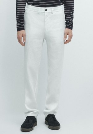 Uomo con pantaloni bianchi a gamba dritta, maglia a maniche lunghe a righe bianche e nere e sneakers nere, in piedi contro uno sfondo neutro.