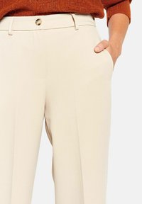 Beige maßgeschneiderte Hose mit glatter Textur, ausgestattet mit Fronttaschen, einem Gürtelschlaufen und einem goldenen Knopfverschluss.