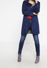Abrigo de ante azul marino con un forro interior gris, que cuenta con un collar y mangas largas. Combinado con una blusa roja, jeans azules y botas cortas grises.