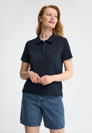 Mujer con pelo rojo que lleva una camisa polo azul marino con un logo rojo en el pecho y pantalones cortos vaqueros azules de longitud media, de pie frente a un fondo blanco.