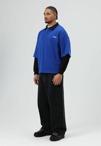 Blauw poloshirt met zwarte kraag, gecombineerd met zwarte broek met wijde pijpen en zwarte schoenen. Het shirt heeft korte mouwen en subtiele branding.