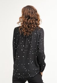 Blouse noire en tissu léger avec un motif d'oiseaux blancs. Manches longues, ourlet arrondi et dos plissé.