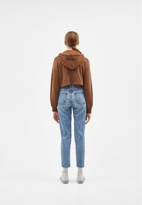 Mujer de pie de espaldas a la cámara, vistiendo un hoodie corto marrón, jeans azules de cintura alta, calcetines blancos y zapatillas blancas.