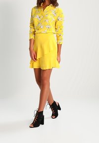 Gelbe Bluse mit Blumenmuster, kombiniert mit einem gelben, gestuften Rock und schwarzen, geschnürten Blockabsätzen. Fokus auf leuchtenden Farben und geschichtetem Design.