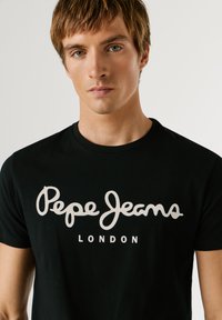 Mladý muž s krátkymi hnedými vlasmi, ktorý má na sebe čierne tričko s bielym logom "Pepe Jeans London" na prednej strane, na jednoduchom pozadí.
