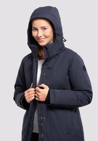 Chaqueta aislante de color azul marino con capucha, cremallera frontal y botones a presión. Presenta una textura suave y puños elásticos. Logo en la manga.