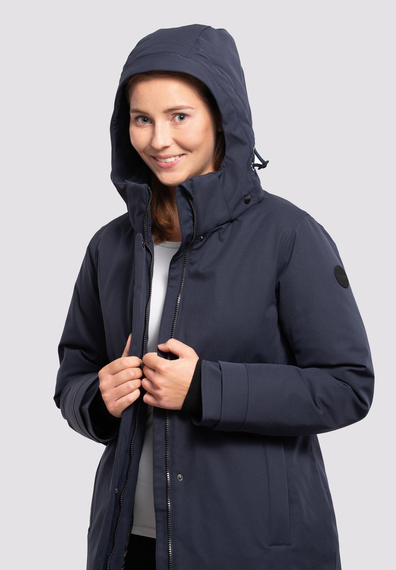 Chaqueta aislante de color azul marino con capucha, cremallera frontal y botones a presión. Presenta una textura suave y puños elásticos. Logo en la manga.