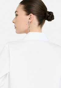 Camicia bianca con colletto, caratterizzata da una trama liscia, con un retro del collo ben visibile e maniche lunghe, abbinata a un orecchino a catena metallica.