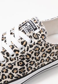 Sapatilhas de canvas com um fundo bege e padrão de leopardo em preto e bege, atacadores brancos e um rótulo preto com texto branco.
