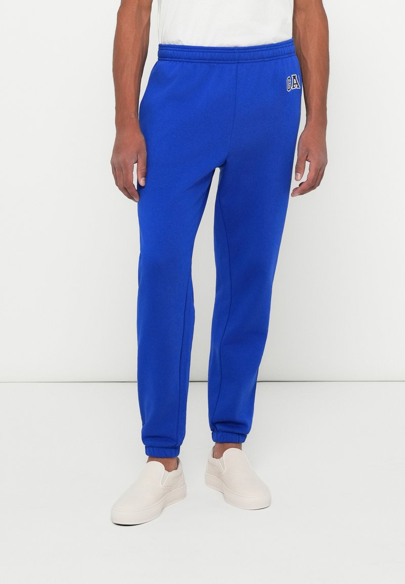 Μπλε αθλητικές φόρμες με ελαστική μέση, μανσέτες στους αστραγάλους και λογότυπο στο αριστερό πόδι. Συνδυάζονται με ανοιχτόχρωμα αθλητικά παπούτσια.