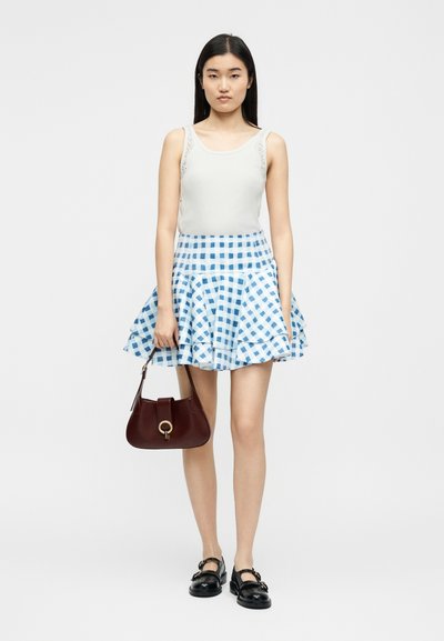 Γυναίκα που στέκεται φορώντας λευκό αμάνικο τοπ, μπλε και άσπρη καρό φούστα, μαύρα παπούτσια και κρατώντας καφέ τσάντα με χρυσό κλείσιμο.