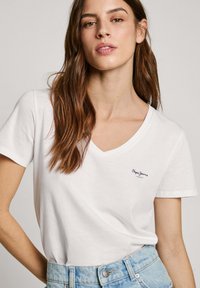 Baltos medvilnės marškinėliai su V formos iškirpte, trumpomis rankovėmis, su maža mėlyna "Pepe Jeans" logo kairėje krūtinės pusėje. Laisvalaikio stiliaus.