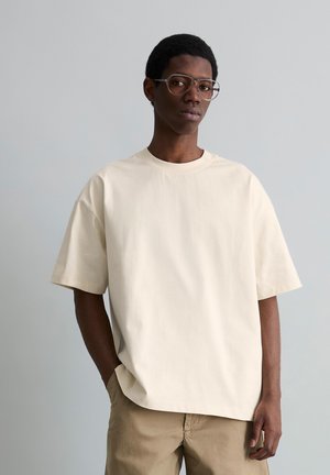 Junger Mann trägt übergroßes cremefarbenes kurzärmeliges T-Shirt und beige Hose, steht mit einer Hand in der Tasche vor grauem Hintergrund.